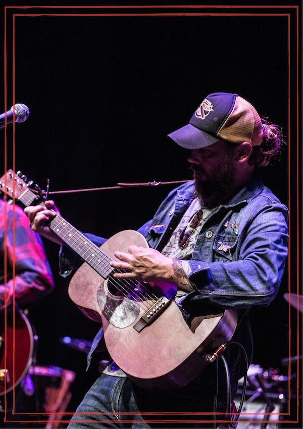 Tony Harrah on stage playing acoustic guitar wearing ball cap