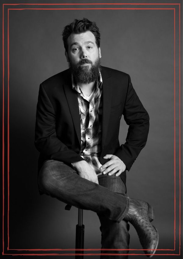 Black & white image of Tony Harrah sitting with legs crossed, wearing dark blazer and boots.